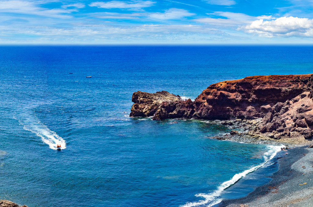 Dunkle Felsküste und schwarzer Kiesstrand auf den Kanarischen Inseln. Ein Boot zieht eine weiße Spur durch das tiefblaue Meer.