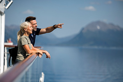 Ein Paar steht an Deck eines Havila Schiffs, blickt auf einen Fjord in Norwegen. Der Mann zeigt mit dem Finger auf die Berge.