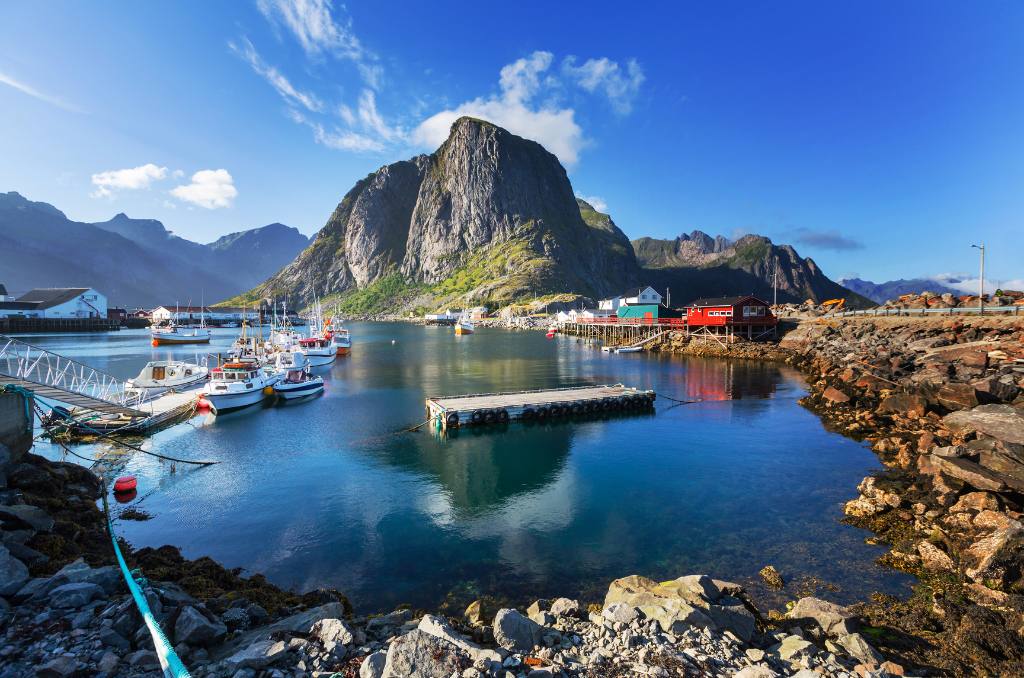 Kleiner Hafen mit mehreren angelegten Booten, umgeben von felsigen Ufern und Häusern auf Stelzen. Im Hintergrund ragt ein markanter, steiler Berg auf, dahinter weitere Gebirgsketten. Der Himmel ist klar und blau mit vereinzelten Wolken, das Wasser spiegelt die Umgebung ruhig wider