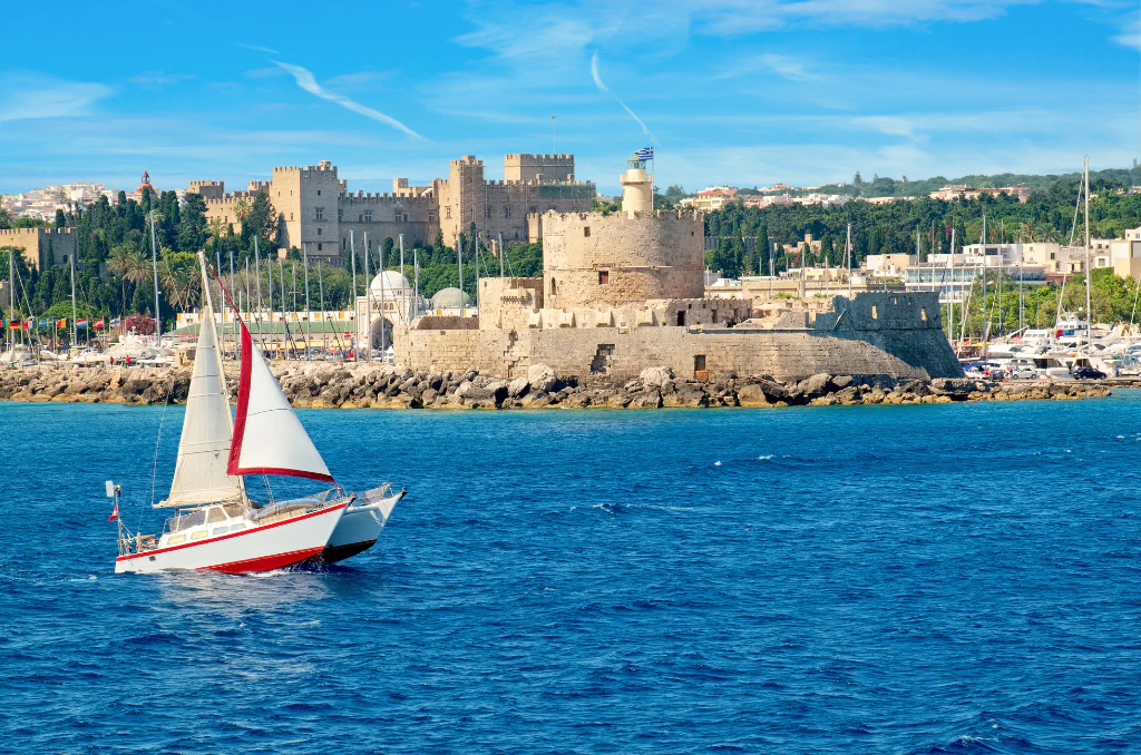 Ein Segelboot mit weißen Segeln und rotem Streifen auf blauem Meer im Vordergrund. Im Hintergrund eine historische Festung mit einem runden Turm und einer griechischen Flagge an einer felsigen Küste. Ein Segelboot mit weißen Segeln und rotem Streifen auf blauem Meer im Vordergrund. Im Hintergrund eine historische Festung mit einem runden Turm und einer griechischen Flagge an einer felsigen Küste.