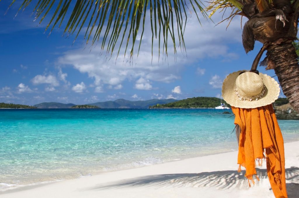 Tropischer Sandstrand mit türkisblauem Meer. Rechts hängt ein Strohhut mit orangefarbenem Tuch an einem Palmenstamm.
